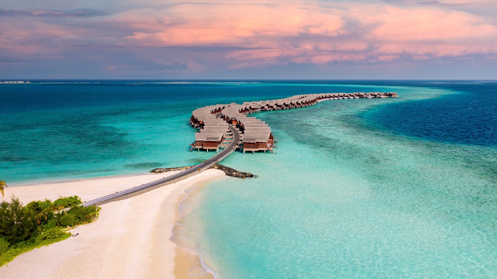 Aerial view of Maldives overwater villas at sunset with a pink sky over a turquoise lagoon.