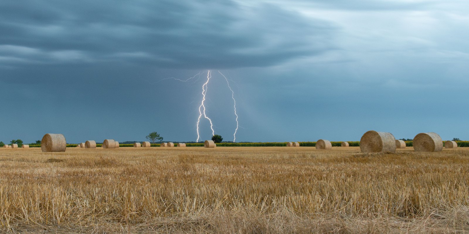 Twin forked lightning strikes on the horizon over a harvested field with hay bales under a storm sky